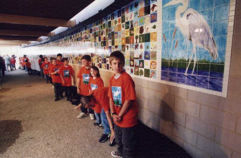 A group of children in red shirts stand along a wall decorated with colorful tile art, including a large blue and white heron, inside a well-lit hallway.