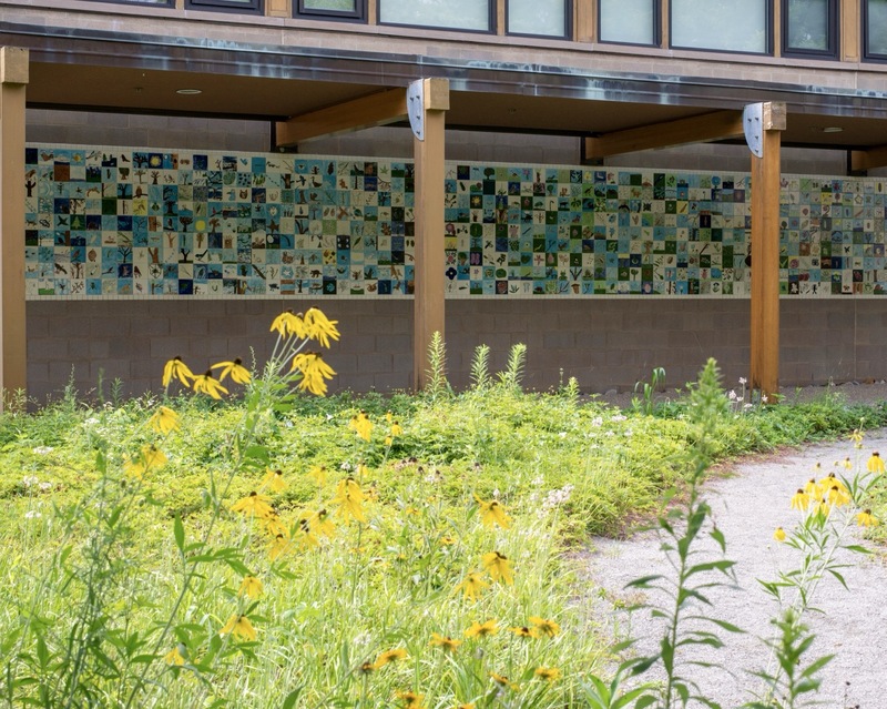 Yellow wildflowers grow beside a paved path in front of a building with a large, colorful tile mural on the wall under a covered walkway supported by wooden posts.