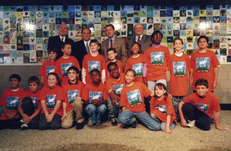 A group of children in matching orange T-shirts poses with four adults in suits in front of a colorful tile mural. The children are smiling, some kneeling in front and others standing behind them.