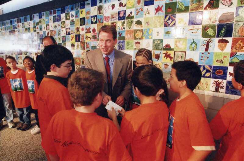 A man in a suit talks to a group of children wearing matching orange shirts in front of a colorful tiled wall decorated with various drawings and designs.