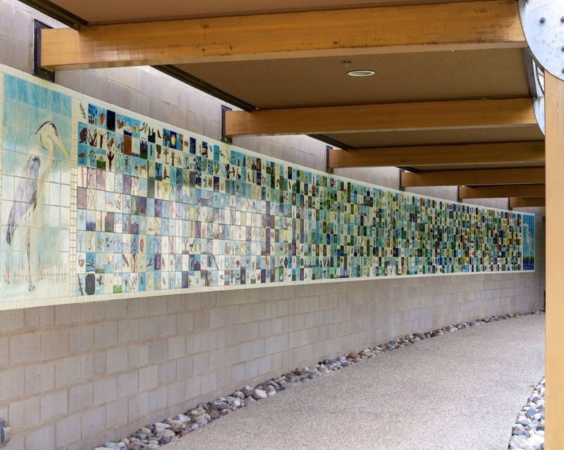 A long, outdoor mural made up of small, colorful tiles depicting various animals and nature scenes, mounted on a brick wall beneath a wooden pergola, with a pathway and rocks alongside.